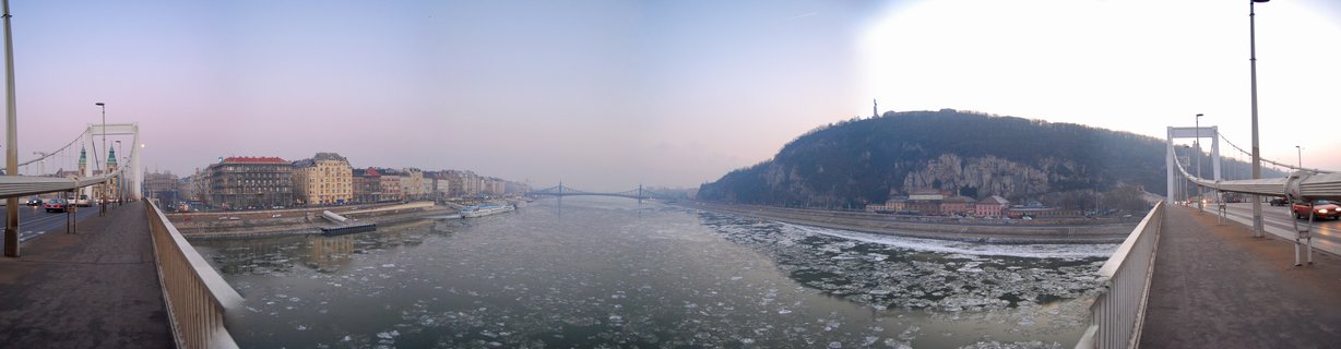 Panoramic winter view of Budapest's Danube River from Elizabeth Bridge at dusk, showing ice on the water, Gellért Hill with Liberty Statue, and city buildings along the riverbank