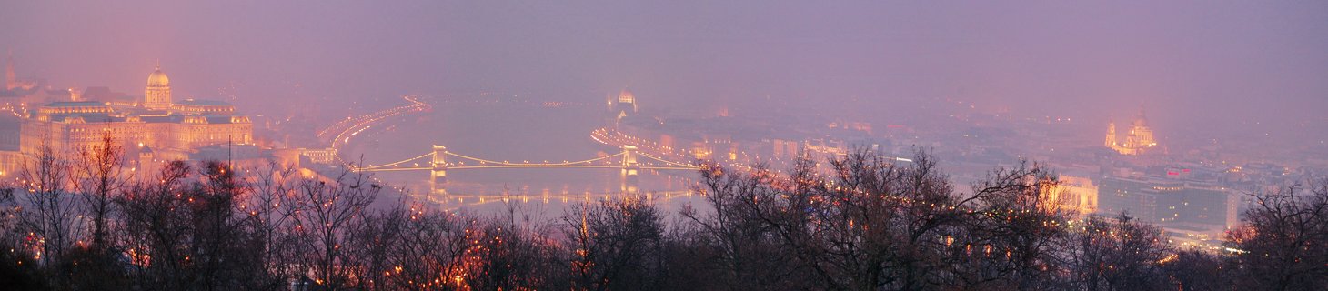 Panoramic view of Budapest at dusk showing the illuminated Chain Bridge over the Danube River with Buda Castle and St. Stephen's Basilica visible through purple twilight haze