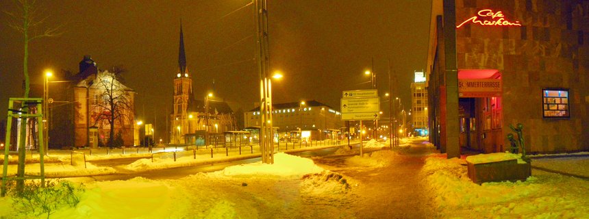 Night photograph of a snow-covered street in Chemnitz, Germany with illuminated buildings, a church spire, streetlights, and visible signage including Café Markoni neon sign
