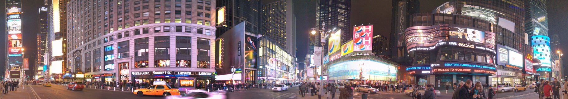 Panoramic nighttime photograph of Times Square showing illuminated billboards, yellow taxis, pedestrians, and tall buildings in New York City