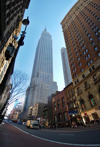 Low-angle street view of the Empire State Building rising between surrounding buildings in Manhattan, with pedestrians, vehicles, and storefronts visible at street level under a clear blue sky
