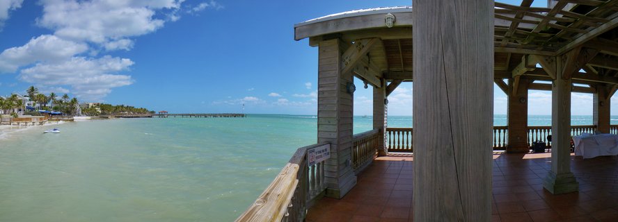 Panoramic view of a Key West beach showing a wooden pier structure on the left, white sand beach with people on lounge chairs, palm trees, resort buildings, and turquoise ocean water under a blue sky with clouds