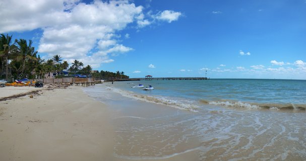 Wide beach view showing sandy shore, turquoise ocean water with two jet skis, a long pier extending into the sea, and palm trees along the coastline under a blue sky with white clouds