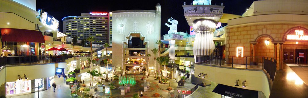 Night view of a multi-level outdoor shopping and entertainment complex with illuminated buildings, palm trees, green fountains, and the Egyptian Theatre facade visible in Hollywood, Los Angeles