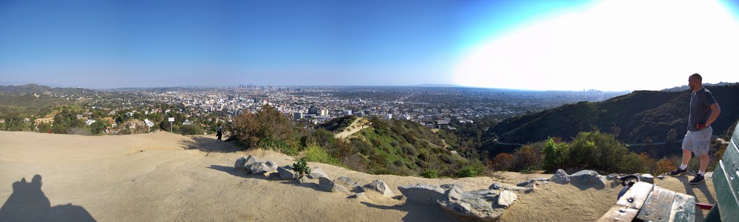 Panoramic photograph showing the Los Angeles cityscape from an elevated hillside viewpoint with downtown skyline visible in the distance under a clear blue sky