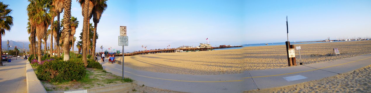 Panoramic view of Stearns Wharf pier extending into the ocean at Santa Barbara beach with palm trees lining a walkway, sandy beach in foreground, and boats visible in the harbor under clear blue sky