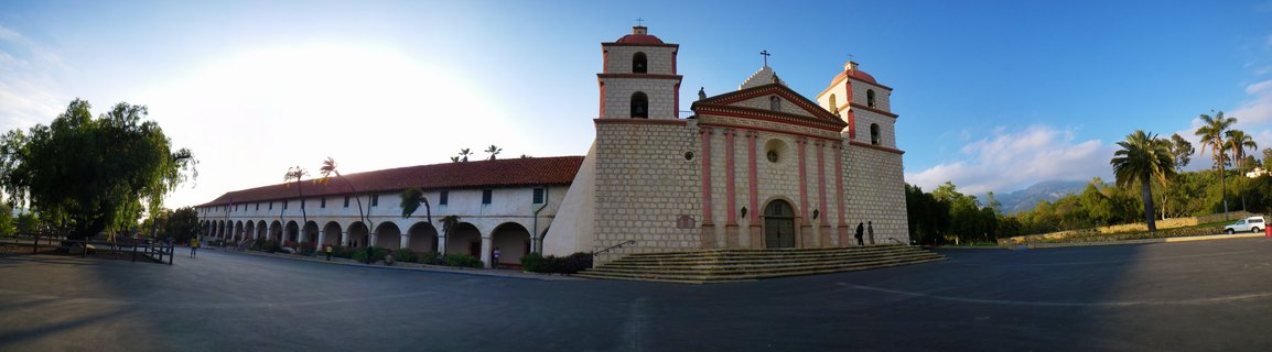 Spanish colonial mission church with twin bell towers, pink sandstone facade, red tile roof, arched colonnade, palm trees, and mountains in background under blue sky