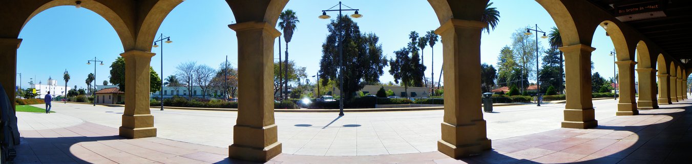 Row of tan stucco arches forming a covered walkway, looking out onto a sunny plaza with palm trees and blue sky in Santa Barbara