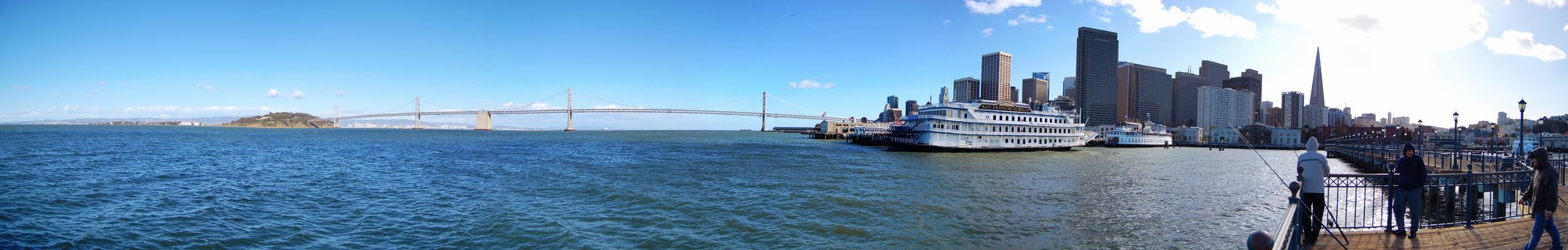 Panoramic photograph of San Francisco waterfront showing the Bay Bridge, city skyline with Transamerica Pyramid, a white ferry boat at the pier, and people fishing from the waterfront promenade with Alcatraz Island visible in the bay