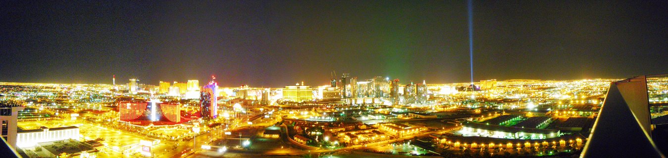 Nighttime panoramic view of Las Vegas Strip with glowing casino hotels, the Luxor pyramid, and a vertical light beam piercing the dark sky.