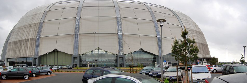 Large white dome-shaped building with curved roof structure, parking lot with cars in foreground, overcast cloudy sky above