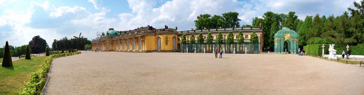 Wide view of yellow Sanssouci Palace with green dome and baroque gardens in Potsdam, Germany, with visitors walking on gravel courtyard under blue sky