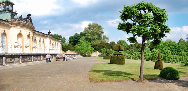 Yellow Rococo palace with green copper dome clock tower, white statues along the facade, manicured topiary bushes on green lawn, gravel pathway, visitors walking, blue sky with clouds