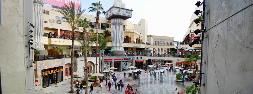 Aerial panoramic view of The Grove shopping center in Los Angeles showing multi-level retail buildings, central fountain display, palm trees, Egyptian-style decorative columns, and shoppers walking on paved walkways