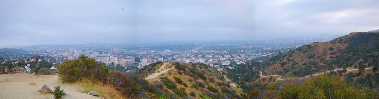 Panoramic photograph showing Los Angeles cityscape from elevated hillside with dry golden-brown terrain and green chaparral vegetation, dirt hiking trails, dense urban development, Downtown LA skyline through atmospheric haze under overcast sky
