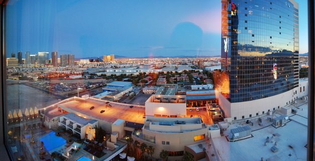Panoramic cityscape of Las Vegas at dusk showing the Hard Rock Hotel tower, swimming pools, parking lots, and the distant Strip skyline under a blue twilight sky