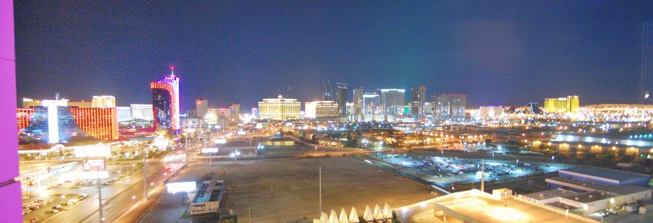 Panoramic night photograph of the Las Vegas Strip showing illuminated casino hotels, neon signs, city lights, highways with traffic, and parking lots under a dark blue sky