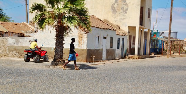 A man in blue shorts walks across a cobblestone street in Cape Verde, passing a palm tree, while another person rides a red ATV. Weathered white buildings with rusted roofs line the background under a clear blue sky.
