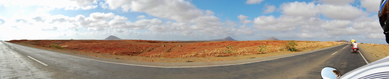Panoramic landscape showing reddish-orange desert terrain with three volcanic mountains in the distance, a paved road in the foreground, and blue sky with white clouds over Sal Island, Cape Verde