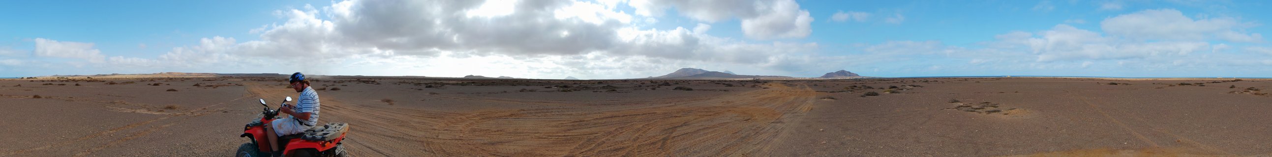 Person riding red ATV across barren reddish-brown desert landscape in Cape Verde with distant mountains and blue sky with white clouds