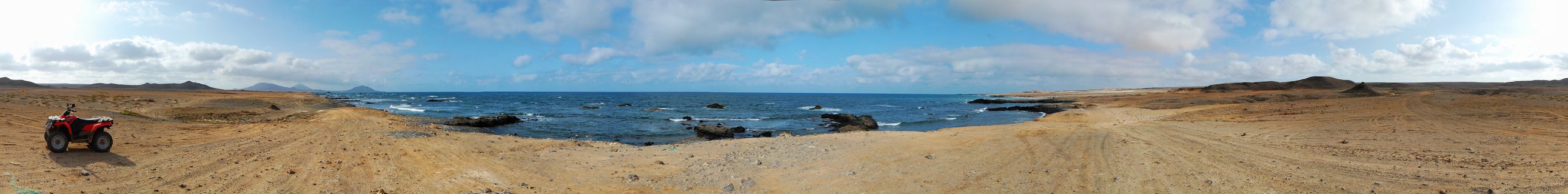 Red ATV vehicle parked on sandy brown coastal terrain with rocky shoreline and blue Atlantic Ocean under cloudy sky in Cape Verde