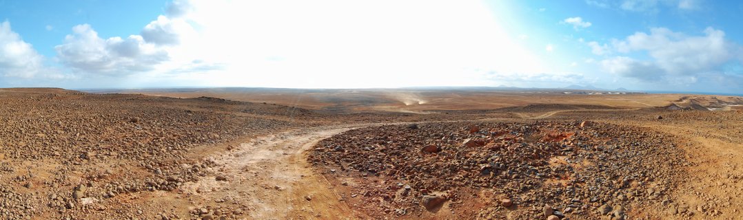 Wide panoramic photograph showing a dirt road curving through rocky desert landscape with reddish-brown volcanic stones covering the ground, bright blue sky with white clouds above, distant horizon showing dust clouds and faint mountains