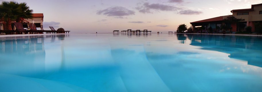 Luxury infinity pool at sunset in Cape Verde resort with turquoise water, palm trees, lounge chairs, and purple-pink sky reflections