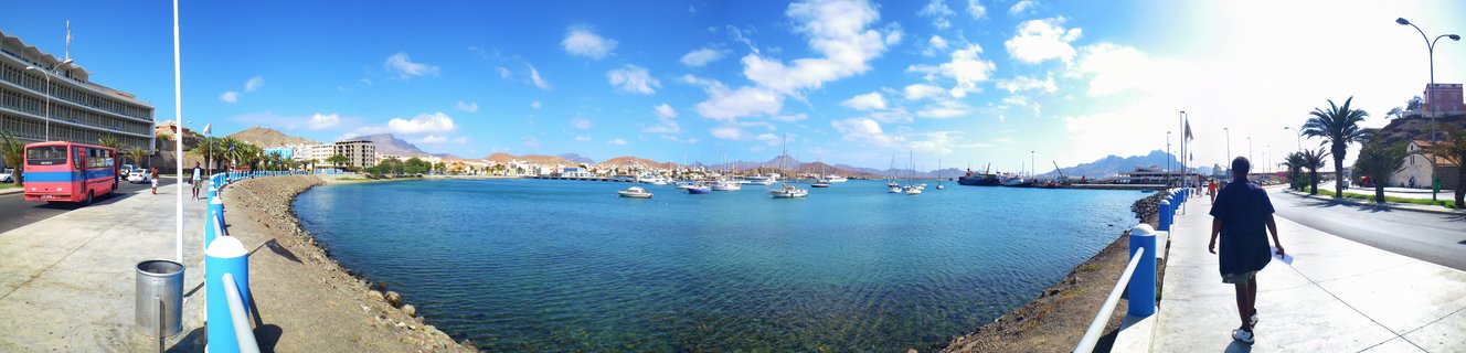 Wide panoramic view of Mindelo harbor in Cape Verde showing turquoise bay water with anchored boats, curved waterfront promenade with blue-white railings, buildings, palm trees, and mountains under blue sky with clouds