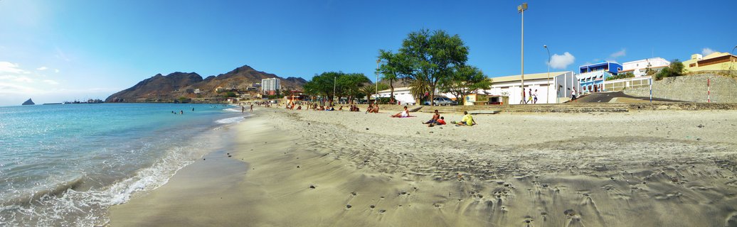 Wide beach scene showing turquoise ocean water meeting sandy shore with people relaxing, trees, white buildings, and mountains in the background under clear blue sky