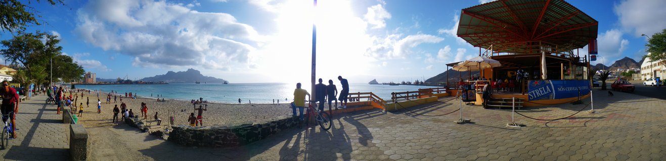 Wide beach scene showing people on sand and paved promenade, ocean with ships, mountains in distance, beach bar structure on right, sunny day with clouds