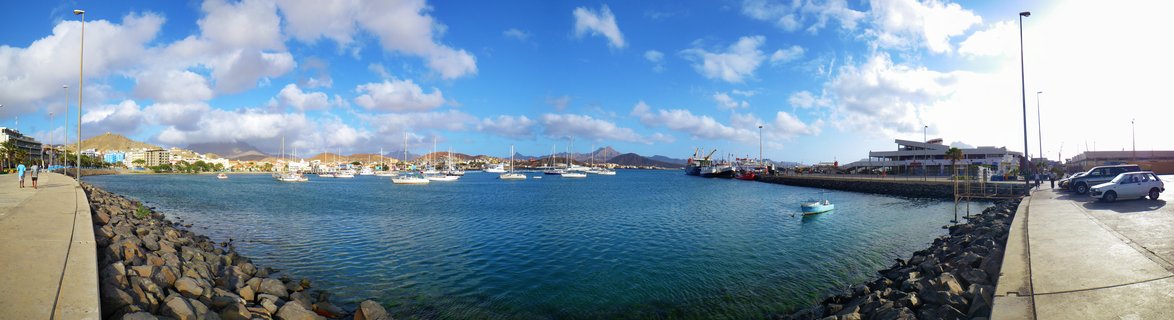 Panoramic harbor scene showing multiple sailboats anchored in blue water with waterfront buildings, rocky shoreline, and mountains in the background under a cloudy blue sky in Cape Verde