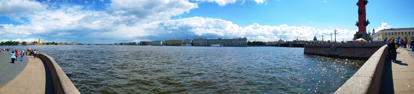 Wide panoramic photograph showing the Neva River in St. Petersburg with a red Rostral Column on the right, historic palace buildings across the water, people walking along stone embankments, and blue sky with white clouds