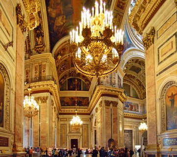 Interior view of an ornate Orthodox cathedral showing large golden chandeliers, painted ceiling frescoes, marble columns, and visitors below