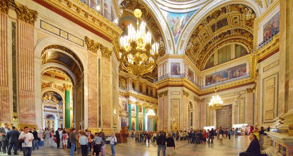 Interior view of ornate cathedral with golden chandeliers, religious frescoes on vaulted ceilings, marble columns, and tourists on polished floor