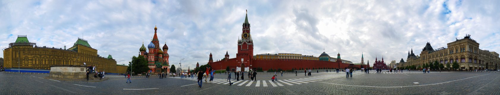 Wide panoramic photograph of Red Square in Moscow showing St. Basil's Cathedral with colorful domes, red Kremlin walls with Spasskaya Tower, historic buildings, and pedestrians on cobblestone plaza under cloudy sky
