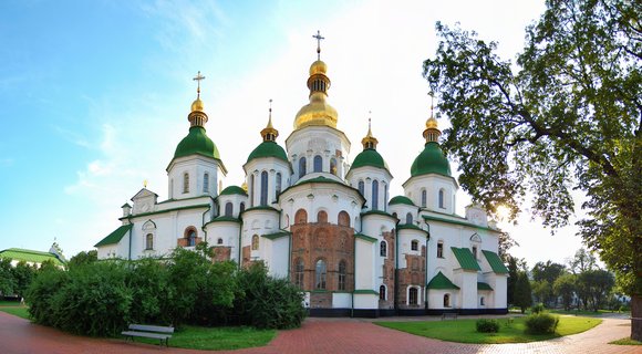 Saint Sophia Cathedral in Kyiv with multiple green and gold onion domes topped with Orthodox crosses, white walls, surrounded by green trees and brick pathways under blue sky