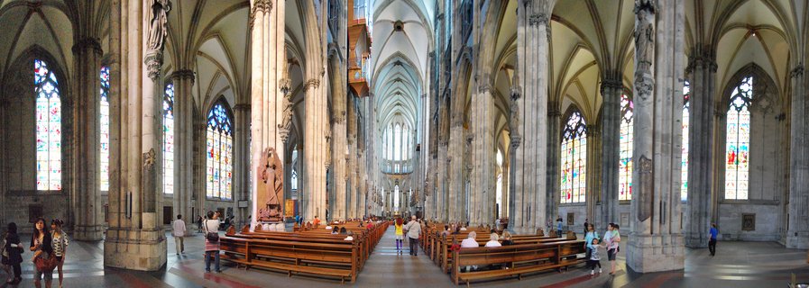Interior panoramic view of Cologne Cathedral showing Gothic stone columns, vaulted ceiling, stained glass windows, wooden pews, and visitors walking through the nave
