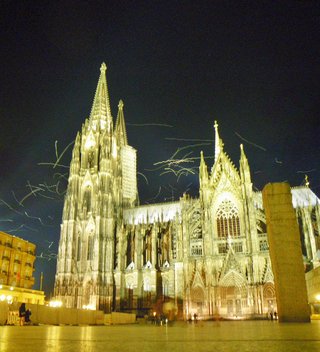 Night photograph of Cologne Cathedral showing illuminated Gothic twin spires and ornate stone facade against dark sky with light trails