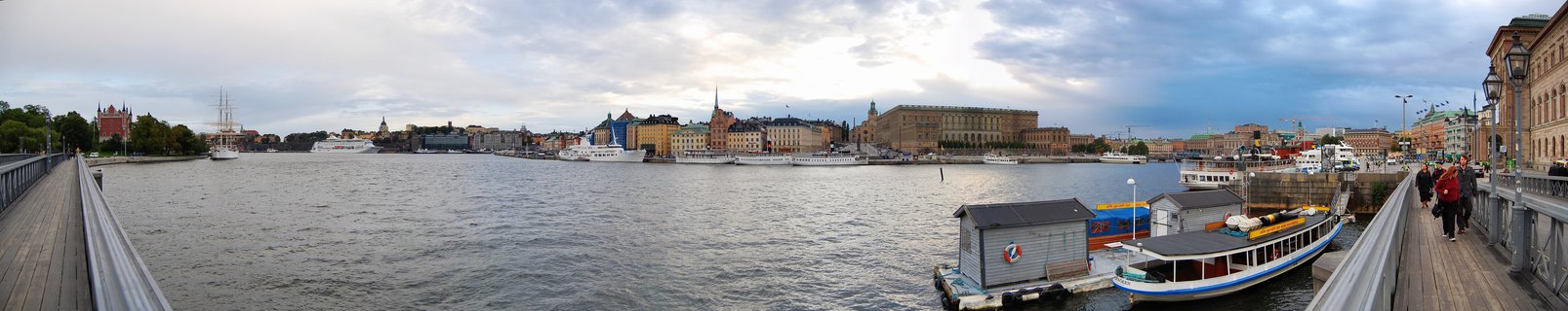 Wide panoramic view of Stockholm waterfront showing Royal Palace, colorful Gamla Stan buildings, boats in harbor, and people walking on bridge under cloudy sky