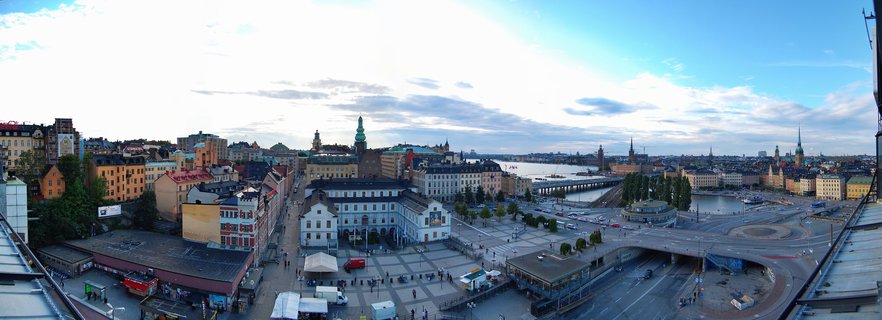 Panoramic cityscape of Stockholm's Old Town showing colorful historic buildings, church spires, the Royal Palace complex, waterfront, bridges, and town square with pedestrians under a partly cloudy sky