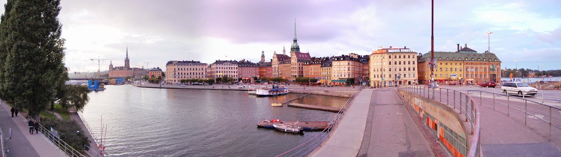Panoramic waterfront view of Stockholm's Old Town with colorful historic buildings, church spires, boats at docks, and pedestrians on promenades under a cloudy evening sky