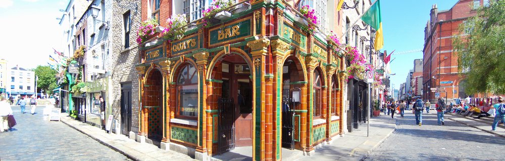 Ornate Victorian pub facade with amber and green terracotta tiles, arched doorways, hanging flower baskets, and BAR signage on cobblestone street in Dublin with pedestrians walking by