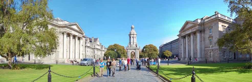 Wide view of Trinity College Dublin's Front Square showing the Campanile bell tower centered between two classical stone buildings with columns, green lawns, and visitors walking on cobblestone paths under blue sky