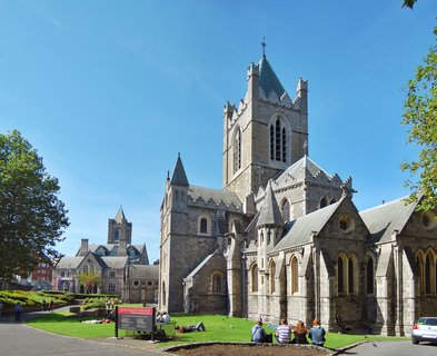 Christ Church Cathedral in Dublin, Ireland showing medieval stone church with tall tower and green copper roof under clear blue sky with people sitting on grass lawn