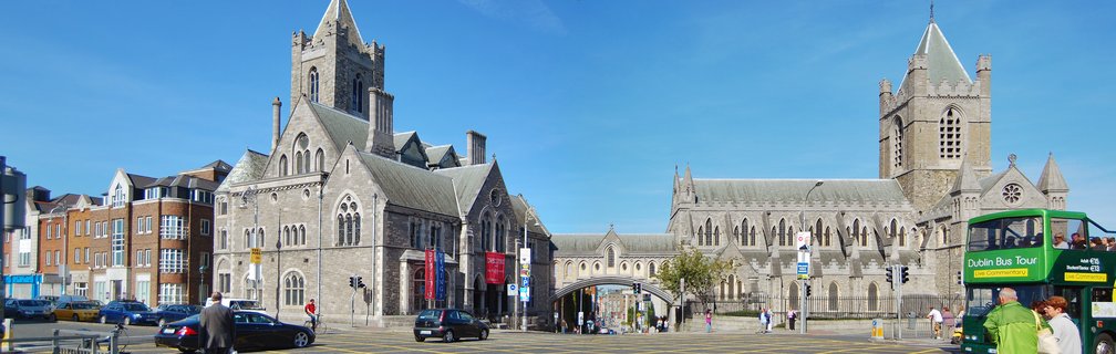 Two medieval stone cathedral buildings with Gothic spires and arched windows under a clear blue sky in Dublin city center, with a green tour bus and pedestrians in the foreground