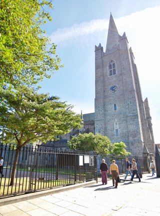 Stone Gothic cathedral with tall pointed spire and clock face, pedestrians walking on sidewalk beside black iron fence, green trees on left, blue sky with clouds