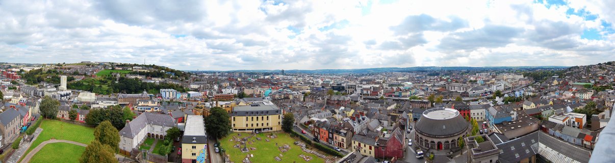 Panoramic aerial view of Cork city showing dense urban buildings, green cemetery areas, historic architecture, and cloudy sky