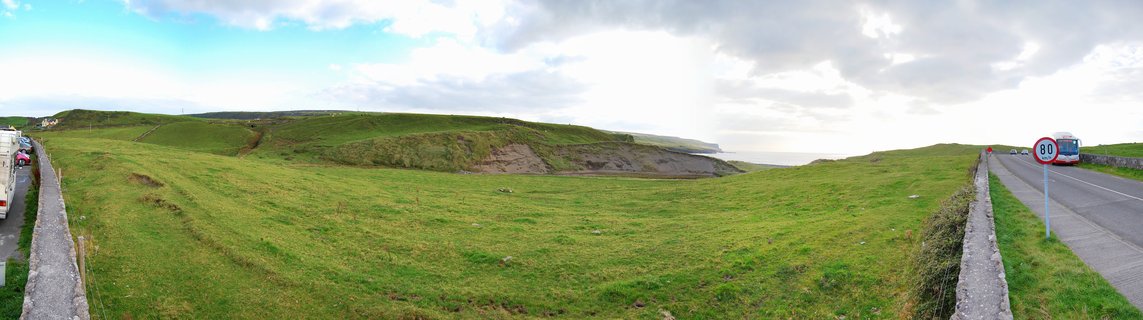 Panoramic landscape showing green rolling hills in rural Ireland with stone walls, a paved road with a red and white bus, and cloudy sky