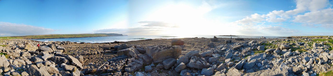 Wide panoramic photograph of a rocky beach with gray limestone boulders in the foreground, blue ocean waves in the middle ground, and green coastal cliffs in the distance under a blue sky with white clouds