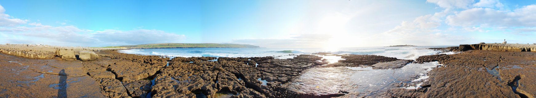 Panoramic view of rocky coastal shoreline with waves breaking, wet limestone formations in foreground, ocean stretching to distant cliffs under blue sky with scattered clouds and bright sun